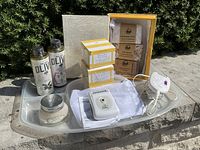 All items laid on a gray tray outdoors showing overall lot contents including L'Occitane cream, shower gels, clay masks, epilator, razor, and soap gift box.