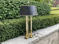 Full view of brass bouillotte style table lamp on a stone ledge with green shrub backdrop