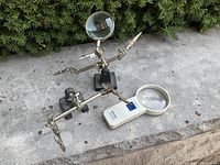 Two metal extra hand magnifiers with round lenses and a battery LED lighted handheld magnifier on outdoor stone surface.