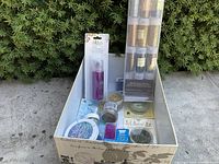 Box containing assorted glitter items outdoors on pavement with shrubs background