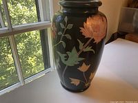 Photo showing side view of ceramic vase with large pink flower, green leaves, and gold outlining on black background.