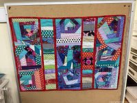 Front view of colorful handmade small quilt with patchwork design pinned to a corkboard.