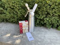 Full view of the Mosa whipped cream dispenser with white handle and nozzle alongside two boxes of cream chargers.