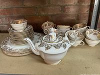 Full view of the Paragon Buckingham teapot with lid, 16 tea cups and saucers stacked beside 4 plates, all white with intricate gold floral and scroll details around edges.
