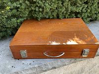 Top of wooden art kit box showing wood grain and shadows from nearby bushes. Box surface has a minor small mark in one corner.