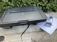 Closed Canon Pixma printer with power cord and two wrapped ink cartridges on white paper.