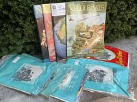 Six petit point needlework kits in original sealed blue packaging displayed with four Needle Arts magazines in the background outdoors.