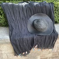 Full view of black crinkled coverup hanging with the black woven wide brim hat placed on it, showing the tassels at the edge.