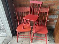 Three red painted wooden chairs; two adult pressback type with decorative carved backs and one smaller child-sized chair stacked on top.