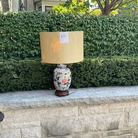 Front angle view of the vintage ceramic lamp with shade, showing floral designs and wooden base placed on stone ledge outdoors.
