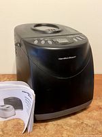 Black Hamilton Beach bread maker with user booklet placed beside it on wooden surface.