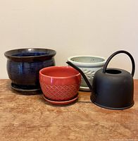 Front view showing three ceramic plant pots of varied colors and designs with a matte black watering can on a wooden surface against a plain wall.