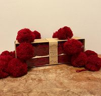 Two sets of red yarn pom pom garlands wrapped and displayed on a wooden surface with several pom poms outside the package showing size and texture.