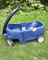 Side view of blue plastic two-seater kids wagon with black handle and white wheels sitting on grass.