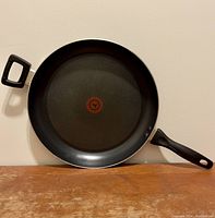 Full view of the black T-fal frying pan, showing the cooking surface and handles on wooden surface against a white background.