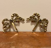 Front view of two solid brass bow-shaped wall hanging hooks symmetrically placed on a wooden surface against a plain wall.