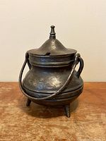 Front view of vintage cast iron pot showing lid with finial handle and side handle attached to loops, standing on three legs.