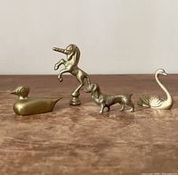 All four brass animal figurines shown in a line on a wooden surface: unicorn, dachshund dog, swan, and duck.