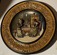 Full view of round terracotta wall charger with ornate frame and deep relief center scene of three women having tea.