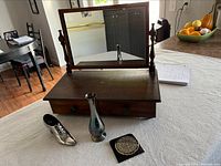 Wooden mirror stand on a tabletop with silver boot figurine, silver plate bud vase, and enamel compact placed in front.