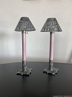 Pair of Victorian candlesticks with silver plate filigree bases, violet candle covers, and perforated silver shades on a black surface against a white wall.