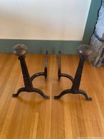 Pair of black iron andirons with brass tops positioned on hardwood floor showing full and side views.