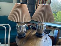 Pair of polished metal vintage lamps placed on a wooden table, showing their overall design and pleated beige lampshades.