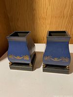 Pair of blue metal urns with gold toile pattern on black bases, showing front view.