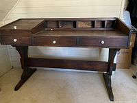 Front view of vintage wood trestle desk showing three front drawers, cubbies on back raised panel, angled lid compartment on left, and sturdy trestle legs.