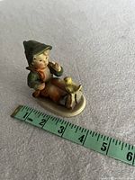 Photo of vintage Hummel figurine sitting with a bird, shown next to measuring tape for size reference about 3 inches long and high.