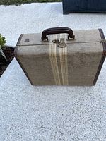 Front angled view of vintage tweed suitcase showing leather handle on top and cream colored horizontal stripes with metal lock.