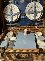 Full view inside the picnic basket showing plates secured with blue leather straps, cutlery attached, thermos, plastic containers, and ceramic mugs and shakers packed inside.