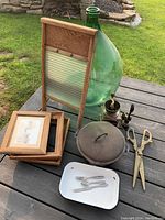 Photo showing the group of antique items including the washboard, picture frames, large green glass demijohn, metal pot with lid, brass pepper grinder, and vintage scissors on a wooden surface outdoors.