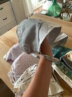 Photo showing a grey bonnet with lace detailing held in hand, various other bonnets and hats visible in background on wooden table.