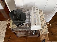 Metal towel rack, black wire hanging basket, covered black plastic wastebasket, white plastic toilet paper holder, and a rolled woven patterned rug.