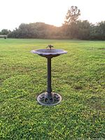 Full view of a metal bird bath standing on grass with the sun setting in the background, showing the overall structure and base details.