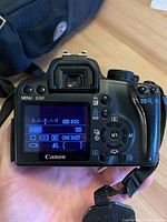 Back view of Canon EOS Rebel XS camera showing LCD screen with settings visible, buttons and dials for operation, and Canon branding on the bottom edge.