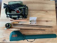 Overview of olive green Cabela's tackle bag, 4-piece fly rod, arbor reel, leather-sheathed fillet knife and fly box on wood surface