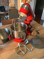 Red Cuisinart stand mixer with attached wire whisk, two stainless steel mixing bowls, flat beater, dough hook, and clear plastic splash guard on wooden table.