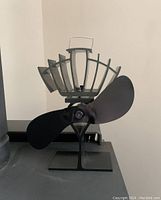 Front and side view of black Ecofan showing blades, metal frame, and sturdy base placed on top of a wood stove.