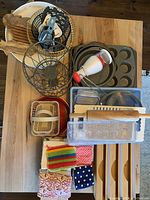 Top-down view of assorted kitchen and baking items arranged on a wooden table including metal baskets, wooden breadboard, muffin tin, hand mixer, plastic organizers, rolling pin, and dishcloths.