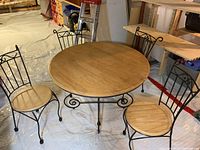 Full view of round wood top bistro table with four wrought iron chairs with wood seats around it on a plastic covered floor.