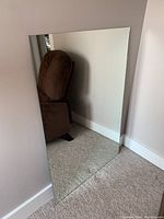 Clear image of large frameless rectangular wall mirror leaning against wall on carpeted floor, reflecting chair and part of room.