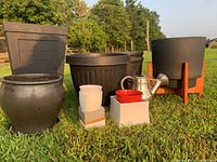Wide shot of lot outdoors on grass, showing various large black and brown plastic and ceramic planters, white and red small pots, and metal watering can.