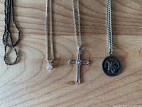 Front view of four necklaces laid flat: a dark chain with no pendant, a delicate chain with diamond pendant, silver cross pendant necklace, and Saint Michael pendant on a thicker chain.
