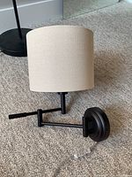 Side view of the plug-in wall sconce lamp lying on carpet, showing beige drum fabric shade, black metal arm, round base, and clear acrylic pull chain.