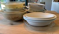 Lots of Pyrex and ceramic bowls and casserole dishes stacked and arranged on wooden table showing different patterns and sizes.