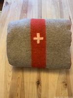 Folded grey wool blanket with red stripe and white Red Cross emblem, showcasing overall design and colors.