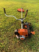 Trimmer angled view showing orange engine housing, bicycle-style handlebar grips, shoulder harness, and straight aluminum shaft