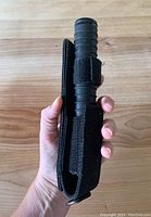 Hand holding the black nylon sheath with tactical baton inside, showing the grip handle and sheath closure.